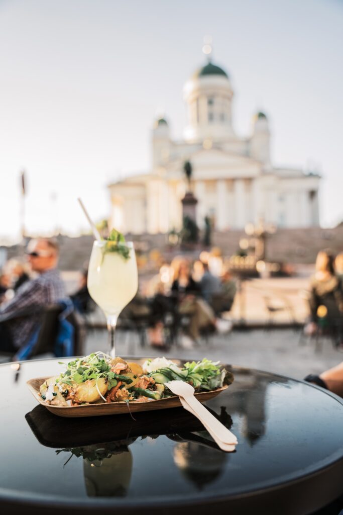 Kuva herkkullisesta pottuannoksesta, jossa on salaattia, perunaa, kalaa ja kastiketta. Ihana herkullinen sitruunainen juoma viinilasissa, koristeltuna. Taustalla näkyy Senaatintori ja Helsingin Tuomionkirkko.Terassi Helsinki